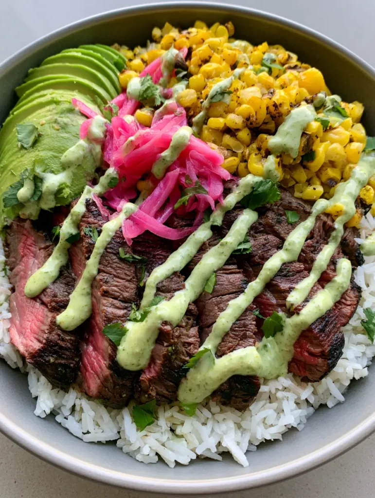 Close-up of charred corn and pale green cilantro cream sauce zig-zagged across a Steak and Avocado Rice Bowl.